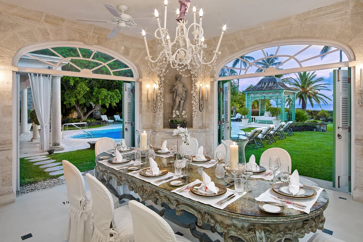 Sophisticated dining room with a vintage-style table set for dinner, elegant white chairs, and open views to a lush garden and pool area under the evening sky in Barbados