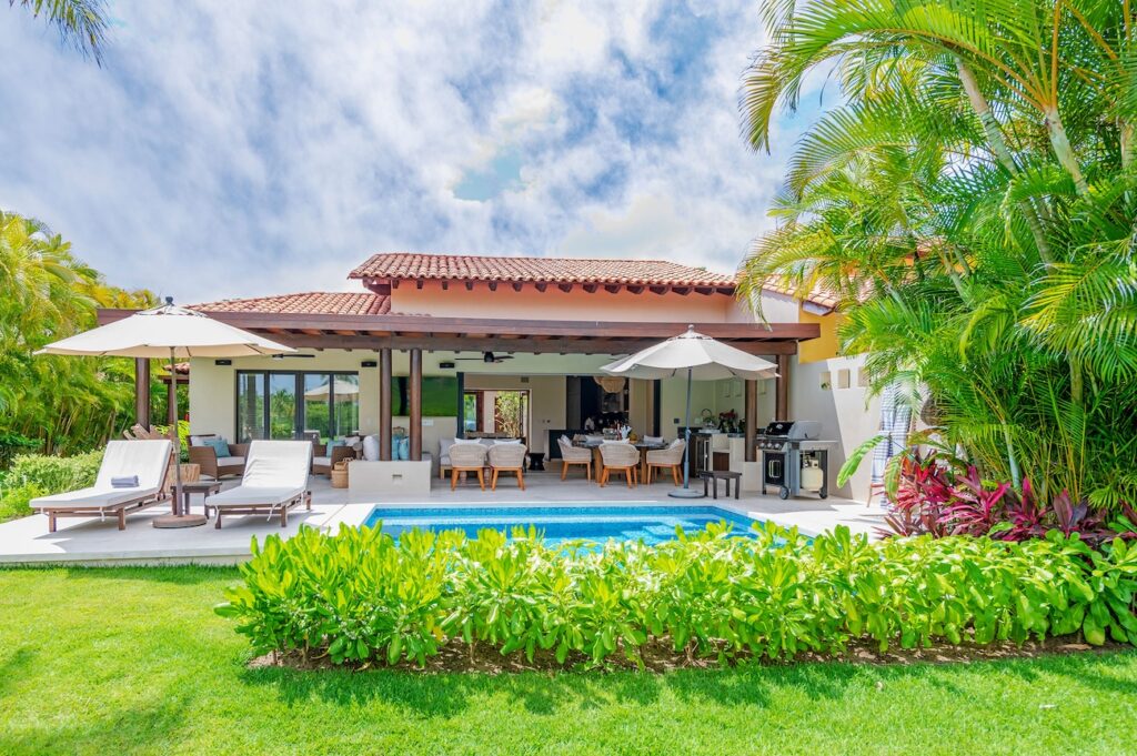Luxurious outdoor patio area of a villa with white lounge chairs under umbrellas, adjacent to a small grass-fringed pool, surrounded by lush tropical vegetation, showcasing a serene living and dining area open to the outdoors
