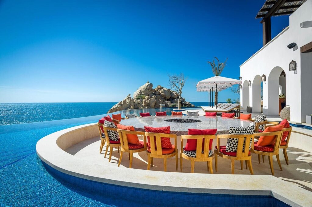 Luxurious outdoor dining area with a circular table surrounded by colorful cushions on seats, located next to an infinity pool with a panoramic view of rocky cliffs and the ocean