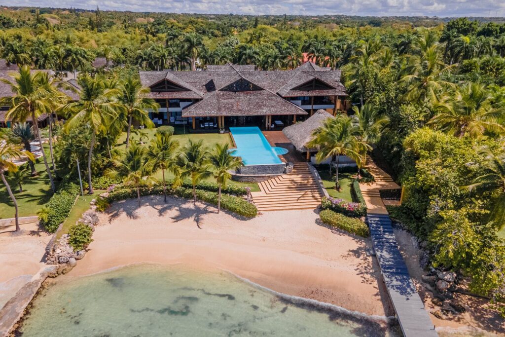 Aerial view of a spacious luxury villa with a thatched roof, large swimming pool, and lush tropical gardens, located directly on a serene sandy beach with clear shallow waters, showcasing the peaceful and exclusive setting in Casa de Campo
