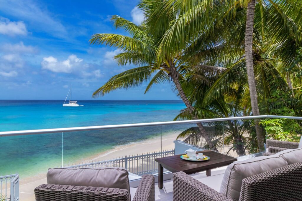 Oceanfront balcony with modern wicker furniture overlooking a serene beach with a yacht sailing in the clear turquoise waters, framed by lush palm trees in Barbados