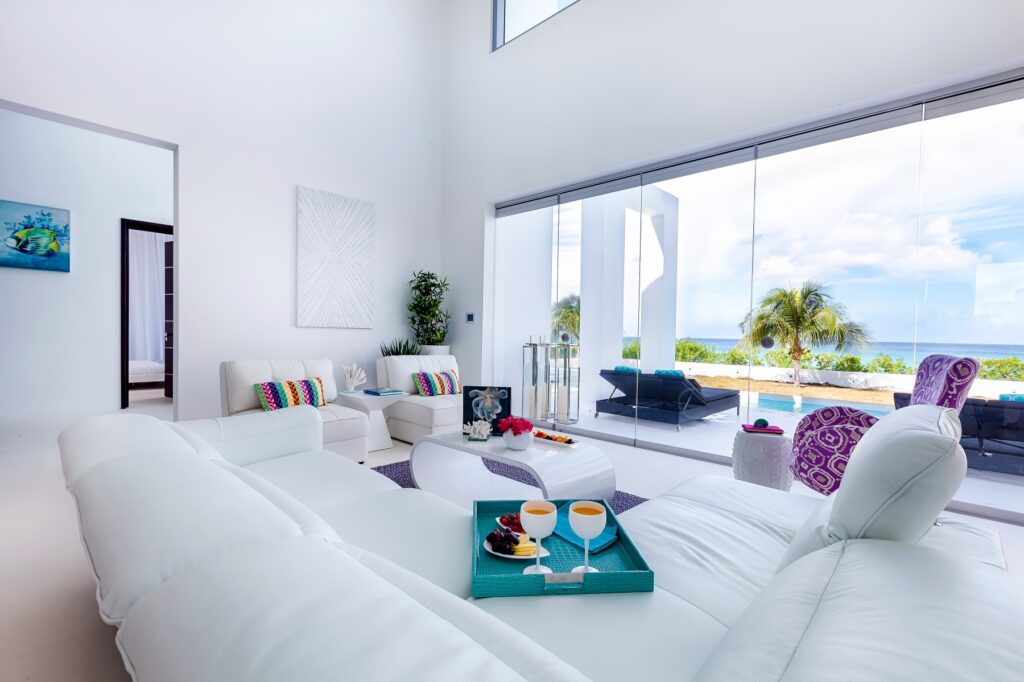 Spacious, brightly lit living room with minimalist white sofas, colorful cushions, and large windows offering panoramic views of a tranquil beach in Anguilla