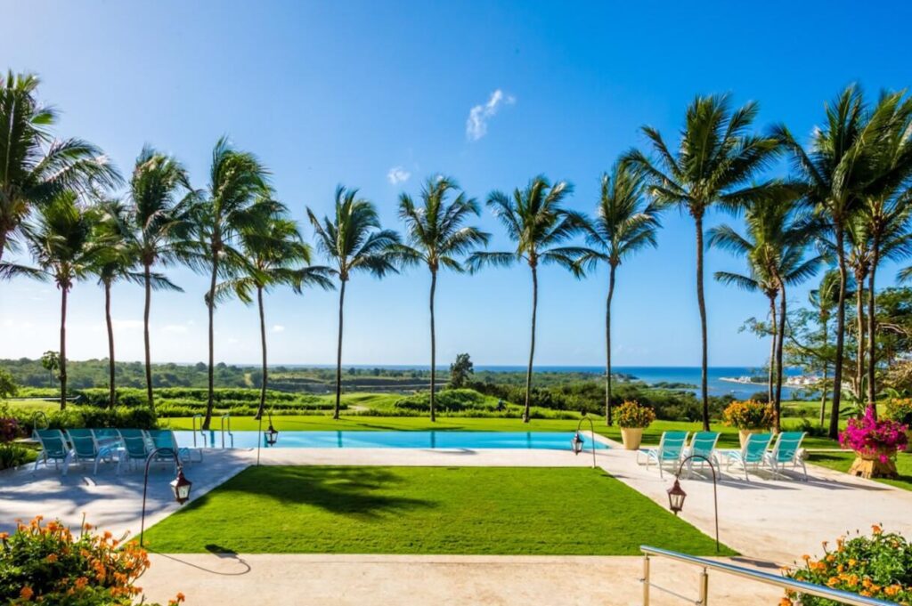 Expansive resort-style pool lined with tall palm trees overlooking a lush golf course and the ocean in the background, under a clear blue sky
