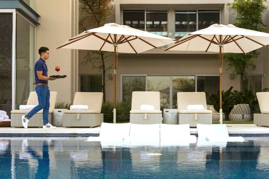 Waiter serving a drink by a modern pool with submerged loungers and large umbrellas, set against a contemporary style residence with lush landscaping in the background