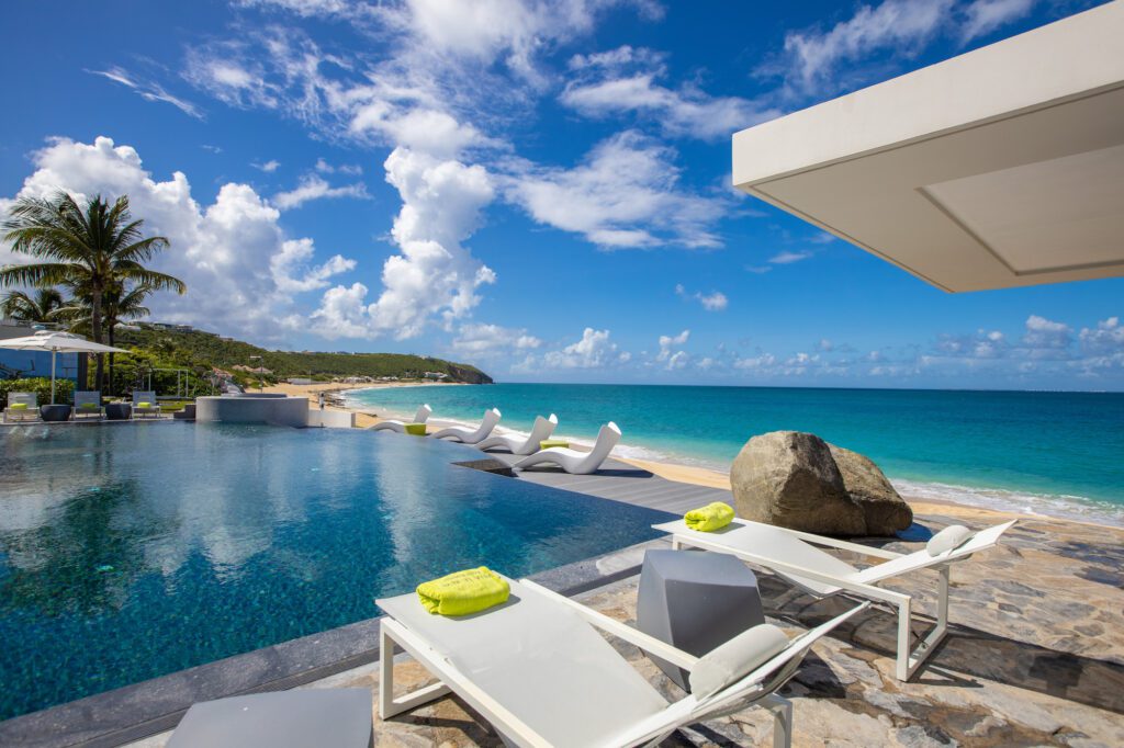 Exquisite poolside lounge with a unique sandy bottom pool surrounded by white cabanas and large rocks, located in a lavish beachfront property in St Martin