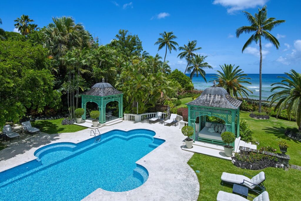 Aerial view of a luxurious estate with a uniquely shaped pool surrounded by lush tropical gardens, gazebo seating areas, and a clear view of the turquoise ocean in Barbados