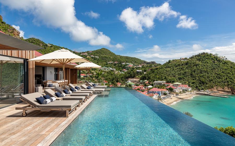 Luxurious Caribbean villa terrace featuring a large infinity pool, wooden deck with sun loungers, and a breathtaking view of a secluded beach and verdant hillsides in St Barts