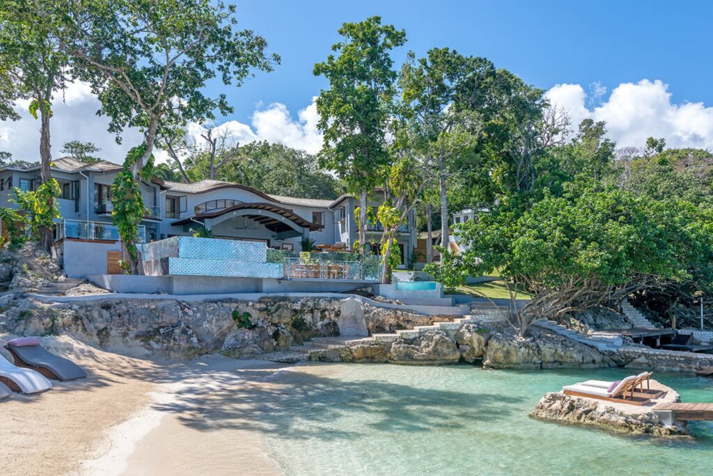 Contemporary beachside villa with unique architectural features and a natural pool, nestled against a backdrop of dense tropical greenery and a private sandy cove in Jamaica