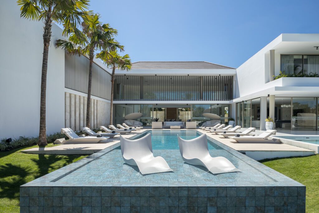 Modern infinity pool with ergonomic white loungers and a stunning view of a minimalist luxury villa, set against a backdrop of palm trees and clear blue skies in Punta Cana