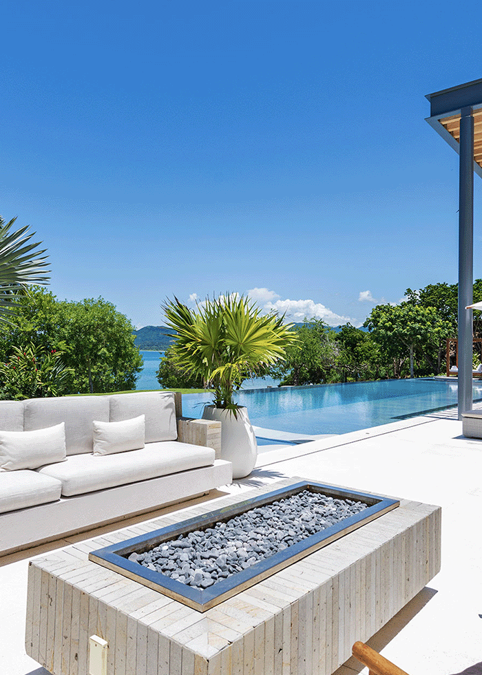 Luxurious outdoor villa with a white sectional sofa and a modern fire pit table, overlooking an infinity pool with a stunning view of the lush landscape and bay in Punta Mita