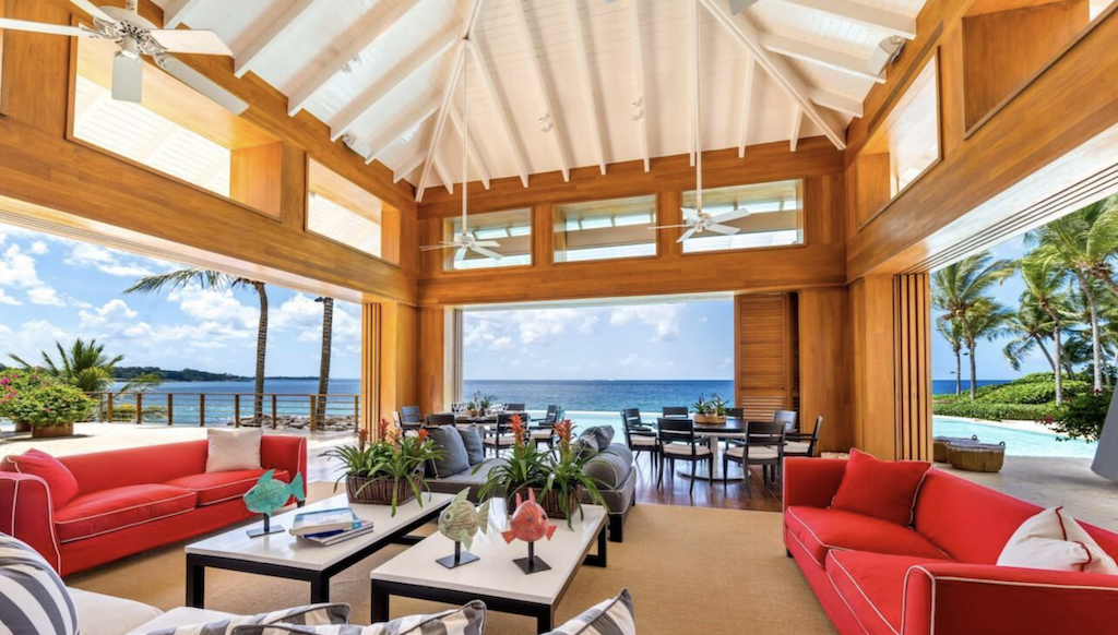 Spacious and airy beachfront living room with high wooden ceilings and panoramic views of the sea, elegantly furnished with vibrant red couches and a large dining area in Casa de Campo
