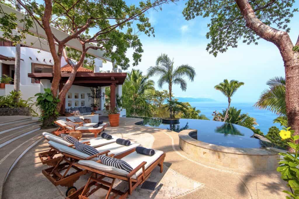 Poolside area with wooden loungers under trees, overlooking an infinity pool and the ocean in Puerto Vallarta