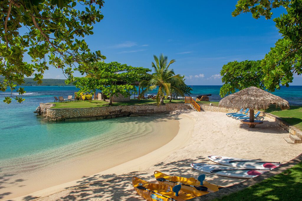 Secluded beachfront property with a grassy lawn leading to a sandy beach, complete with kayaks and shaded seating areas under tropical trees, facing a calm blue sea in Fortlands