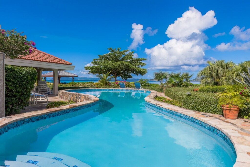 Inviting curved pool with a covered patio and outdoor furniture in a lush garden setting by the sea, featuring panoramic views and vibrant tropical flora in Virgin Gorda