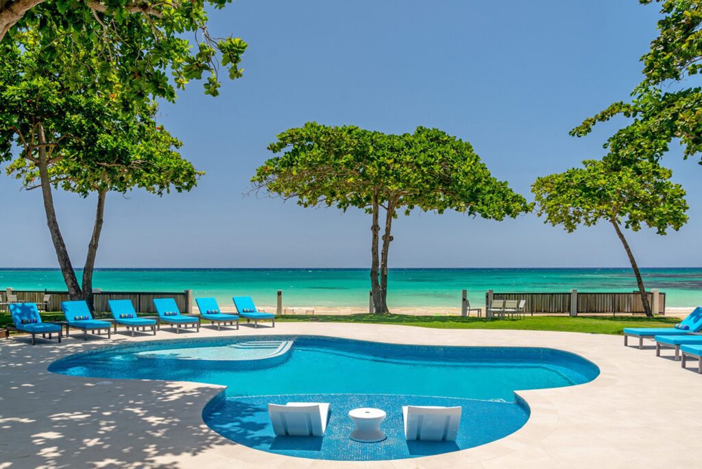 Idyllic beachfront pool area flanked by large shady trees, with loungers overlooking a vibrant turquoise sea, epitomizing luxury tropical living in Jamaica